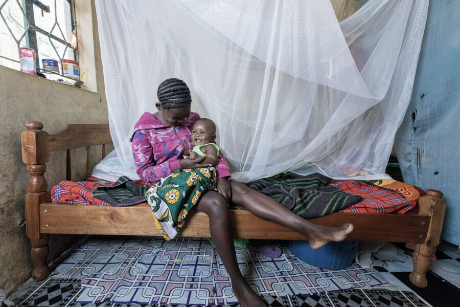 Mother and child sitting on a bed in front of a mosquito net in Uganda.